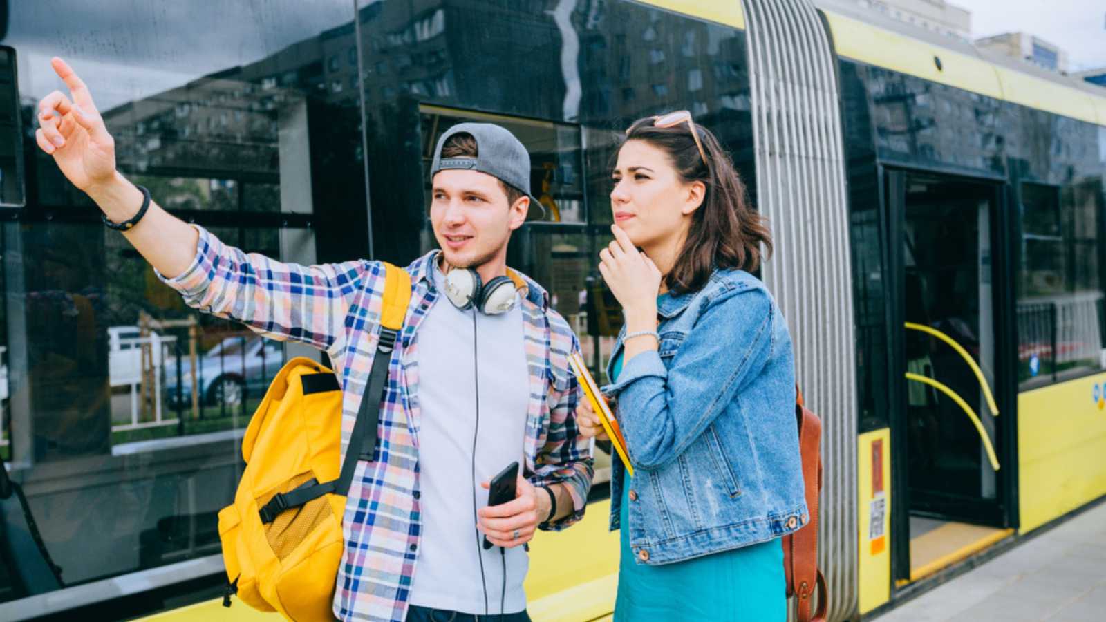 Stylish female traveler with backpack and smartphone in hand asking for help man with backpack at tram stop. Young woman tourist asking for directions and help from local people in tram stop outdoor.