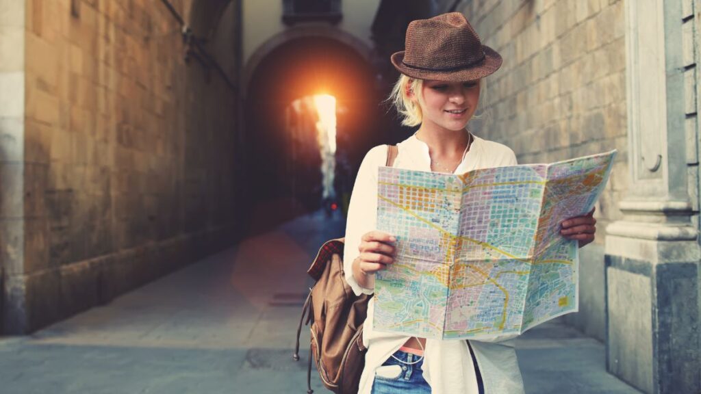 Cheerful woman wanderer with trendy look searching direction on location map while traveling abroad in summer, happy female tourist searching road to hotel on atlas in a foreign city during vacation