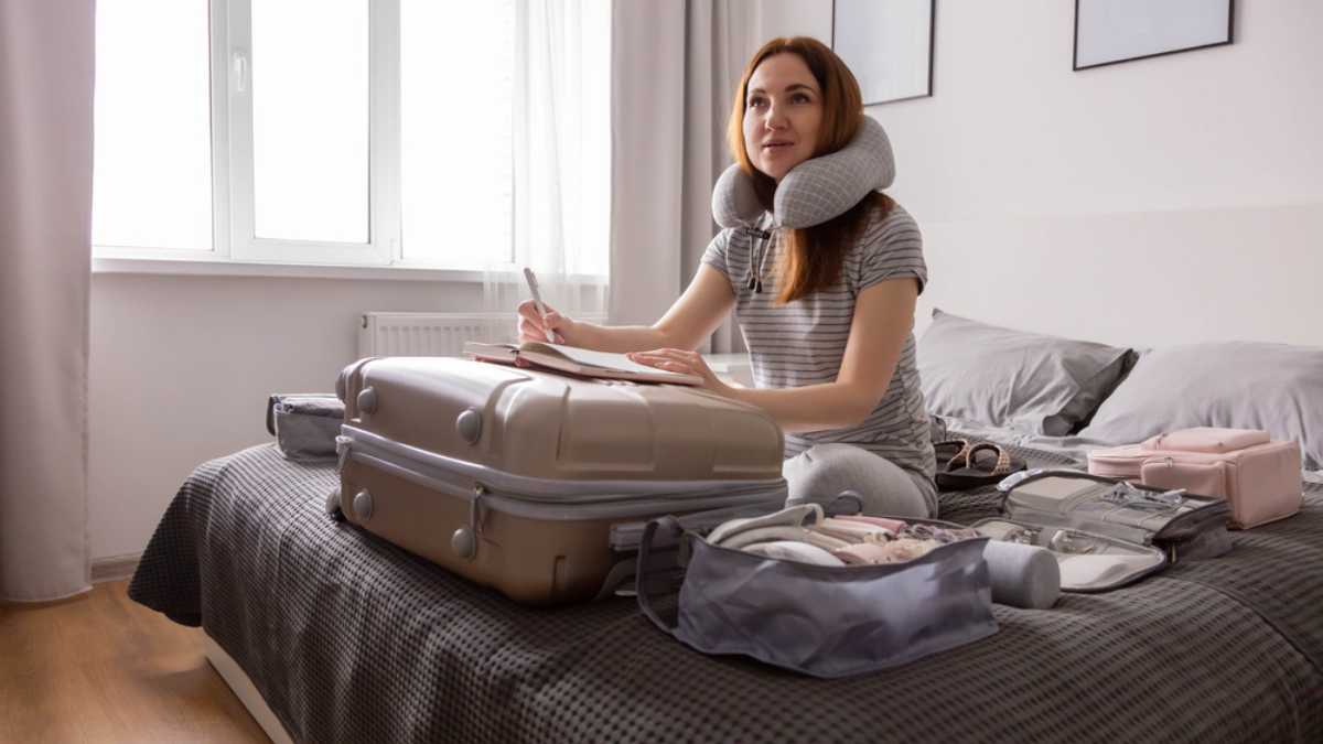 Smiling tourist woman packing suitcase to vacation writing paper list sitting on bed at home. Female getting ready to travel trip organization things storage in comfy cases bags use konmari method