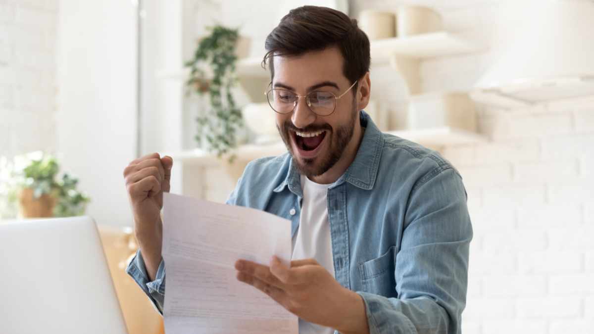 Excited euphoric happy young man holding reading paper postal mail letter amazed overjoyed by good news, got new job celebrate taxes refund receive salary payment loan approval sit at home table