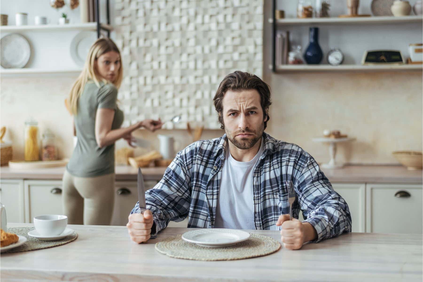 Angry-hungry-dissatisfied-millennial-caucasian-man-with-stubble-ignoring-woman-and-wait-for-food-in-kitchen-interior