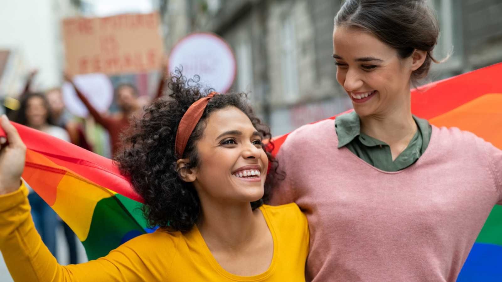 Young women on street enjoying holding gay pride flag during protest. Smiling multiethnic women enjoying during march on street for lgbt rights. Diversity, tolerance and gender identity concept.