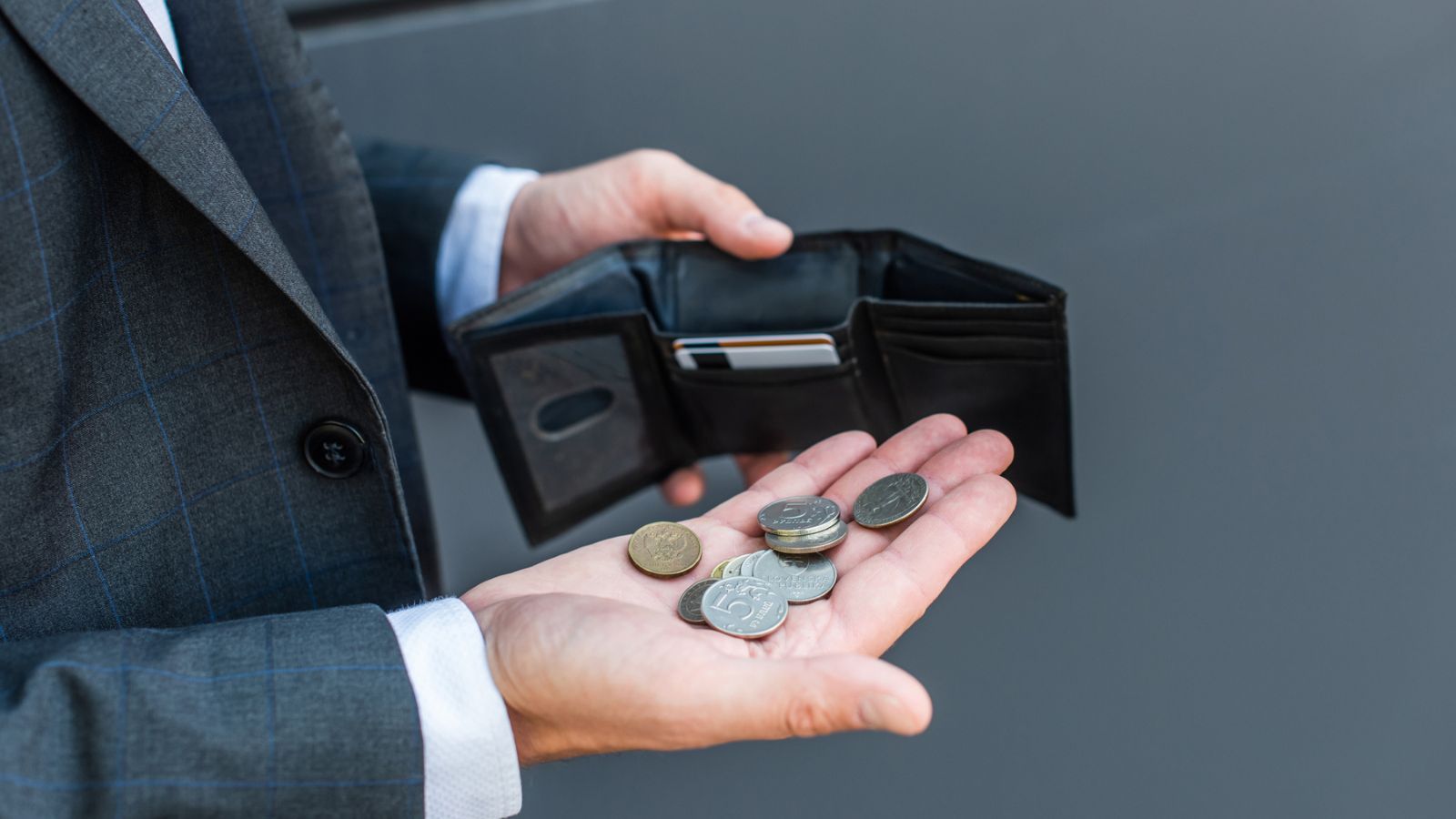 Cropped view of businessman with coins on palm, holding empty wallet with credit cards on grey textured background
